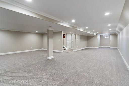 Finished basement with gray carpet, recessed lighting, and support columns.